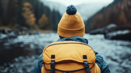 A traveler wearing a dark blue jacket and yellow backpack stands by the flowing river, facing away, with misty forested mountains and autumn foliage in the background.