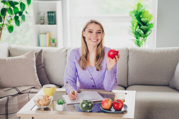 Photo of lovely cheerful girl sit couch table write notebook eat apple dressed violet garment spend...