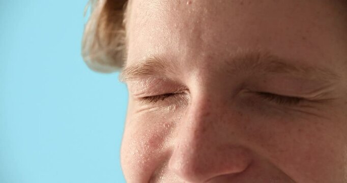 Close up portrait of teenage boy with blue eyes squinting and smiling while applying moisturizing mist to skin