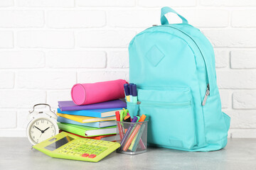 Backpack with different school stationery and alarm clock on light grey table near brick wall