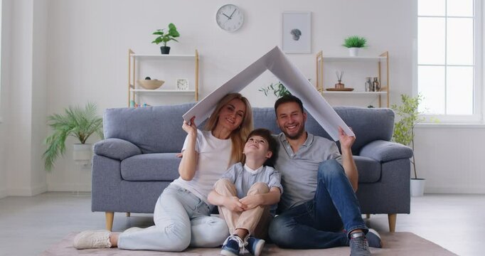 Happy young smiling parents mother, father with child boy sitting on sofa at home holding house roof above. Insurance, mortgage and family protection concept. 4k video. Slow motion video.
