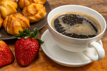 Cup of coffee, fresh strawberries for tasty snack over wooden and profiterole background.