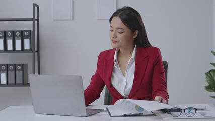 Businesswoman in a red suit sits at her desk, working on a laptop while analyzing financial reports. Focused and determined, she reviews graphs and charts in a modern office setting