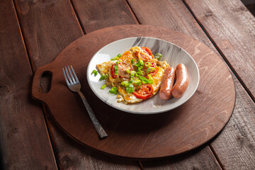 Eggs with tomatoes and fried sausage in a gray plate on a round wooden stand nearby fork