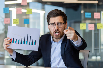 Angry businessman engages in video call, holds chart indicating displeasure during office...