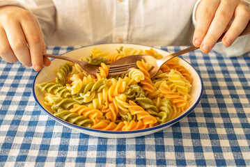 Tricolor pasta for kid's lunch. Plate of pasta and cutlery in the hands of a child.