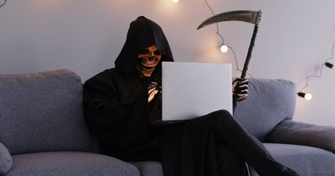 Horrific hacker, Halloween Death man with laptop online, bad horrible fraud looking for new victim, Grim Reaper cosplay costume with scythe, scary man black hooded cape, skull face mask, spooky chat
