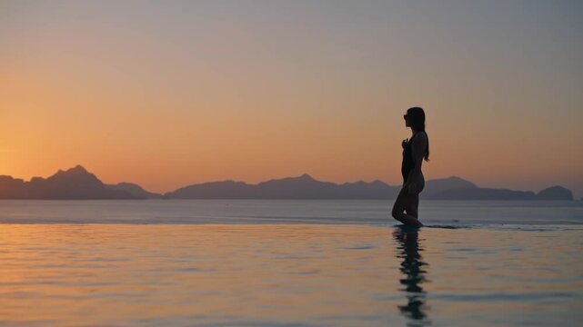 Lifestyle travel video of silhouette woman in swimwear walking on the edge of luxury infinity pool, with golden sunset view reflected in the water, tropical summer vacation 