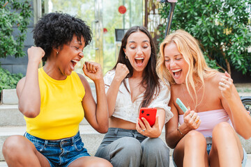 Group of girls looking at smart phone and celebrating winning an online bet. Three succeed young women shouting, cheering, and raising their fists in victory while looking at a cellphone. Achievement