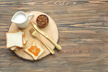 Board of tasty toasts with apricot jam and milk in pitcher on brown wooden background