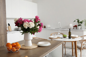 Vase with blooming peony flowers and fruit basket on wooden island table in light kitchen