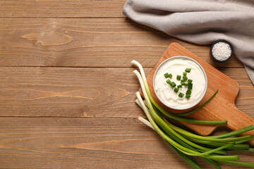 Board and bowl of tasty sour cream with green onion on wooden background