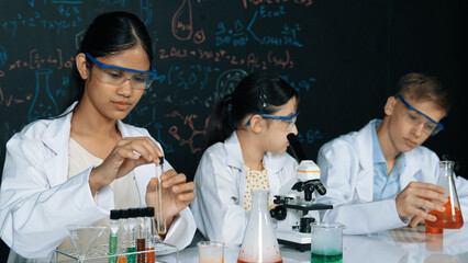 Cute girl looking under microscope while student doing experiment at blackboard with theory...