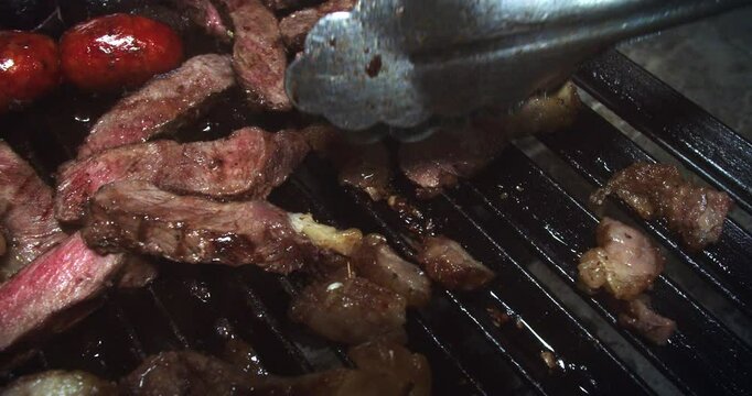 Beautiful slow motion shot of sliced pieces of meat being flipped with chef-operated tongs to cook both sides of the meat on a grill in a restaurant kitchen.