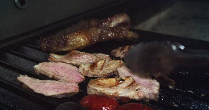 Beautiful slow motion shot of a chef flipping pieces and slices of meat on the rotisserie using large tongs to cook the sides of the meat and sausages inside a kitchen.