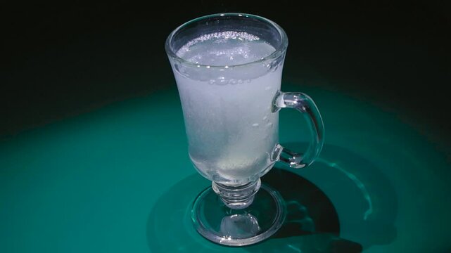 Footage shows effervescent tablet dissolving in glass of water in 4K. Video captures dropping of a vitamin tablet into the water, it starts to fizz and bubble while liquid slowly becomes yellow.
