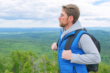 A man in a blue tank top and gray turtleneck stands on a mountain top with a backpack. Close up.