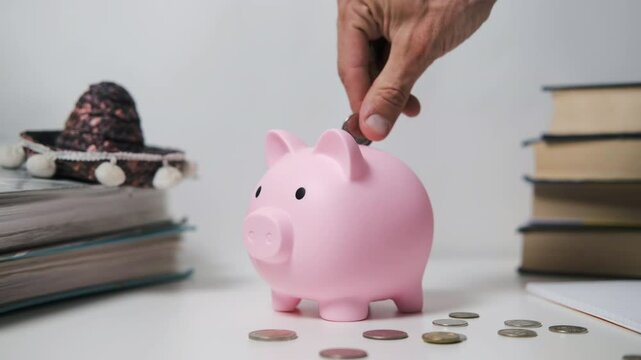 A hand places a coin into a pink piggy bank on a white table, surrounded by scattered coins and a crumpled dollar bill. Savings, financial security and budgeting concept