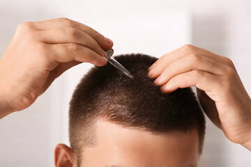 Young man using serum for hair growth in bathroom, closeup. Hair loss concept