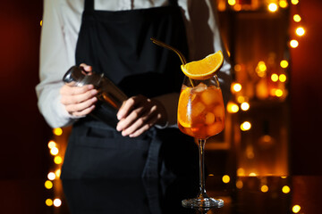 Female bartender with glass of Aperol spritz cocktail on table against blurred lights