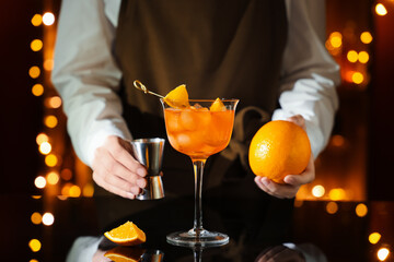 Female bartender with glass of Aperol spritz cocktail on table against blurred lights