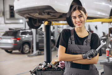 Smiling young woman mechanic standing beneath lifted car, holding tools and looking at camera in...