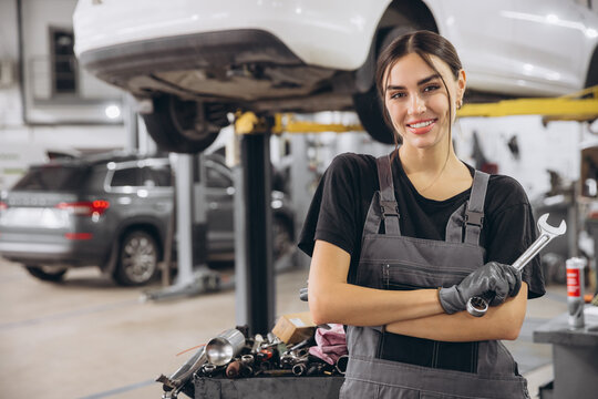 Smiling young woman mechanic standing beneath lifted car, holding tools and looking at camera in car service garage