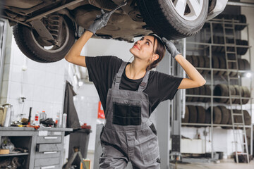 Caucasian mechanic woman fixing underneath car in auto repair shop, female worker in grey uniform doing vehicle maintenance in repair garage.