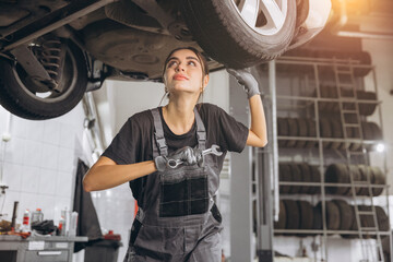 Caucasian mechanic woman fixing underneath car in auto repair shop, female worker in grey uniform doing vehicle maintenance in repair garage.