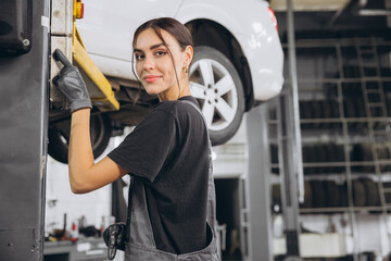 Caucasianl mechanic woman fixing underneath car in auto repair shop, female worker in grey uniform lifts car on auto lift in repair garage.