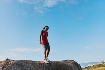 A confident woman in a red outfit stands on a large rock against a clear blue sky, showcasing strength and determination while enjoying nature