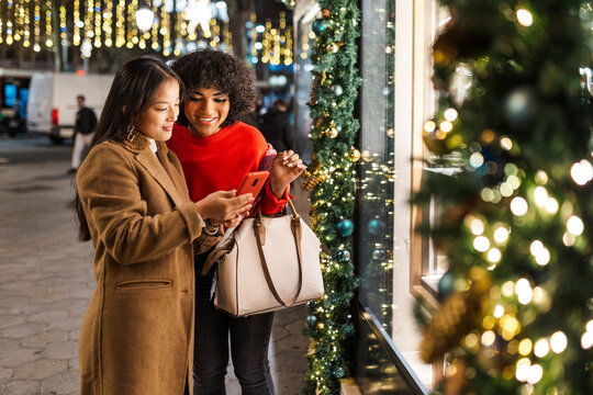 Two young women using smartphone in christmas decorated city at night - Powered by Adobe