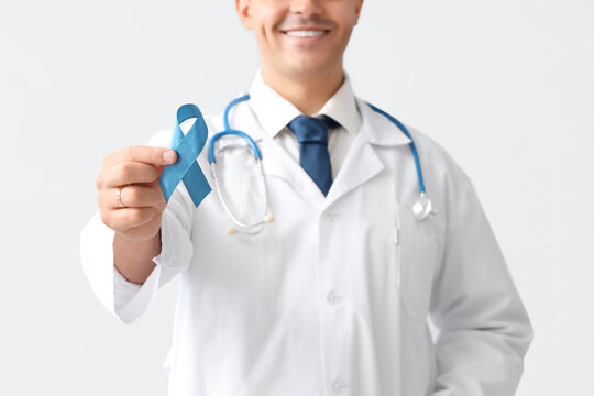 Male doctor with blue ribbon on light background, closeup. Prostate cancer awareness concept