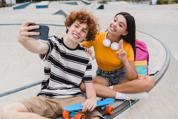 Couple of teenagers classmates students high school pupils friends boy girl bloggers in casual with skateboards bags taking selfie shot holding smart mobile phone having fun sitting on skate park ramp © InsideCreativeHouse