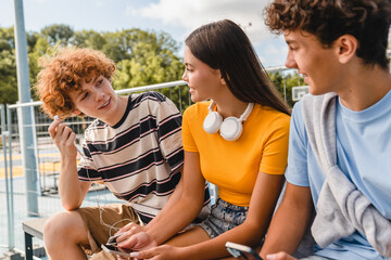Group of smiling friends teenagers high school pupils college students sitting outside in sport yard court using different gadgets listening music having fun hanging out spending time together