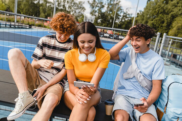Group of smiling friends teenagers high school pupils college students watching funny video laughing out having fun using gadgets internet phones handing out in sport court yard outside