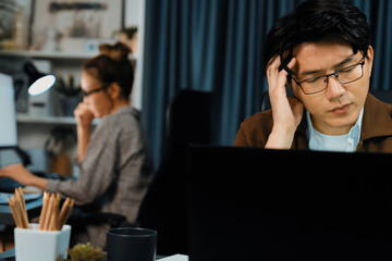Businessman with stressful face working on laptop waiting email while coworker trading stock exchange investment at late night time at modern office, working on startup company project job. Infobahn.