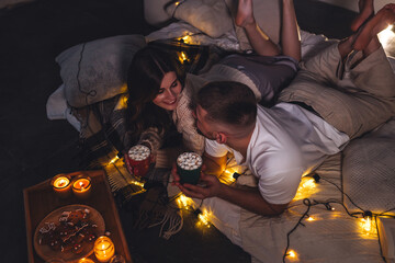 Romantic surprise for girlfriend or boyfriend. Bedroom prepared for watching old movies with popcorn, decorated with lights and candles. Cozy home Christmas atmosphere, hot chocolate with marshmallow