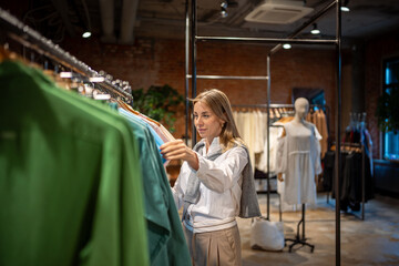 Engrossed, relaxed woman selects items on clothing rack in mall store. Shopping spree for shopaholic, hobby in choosing clothes and items at discount. Consumerism, thrift store, flea market purchase