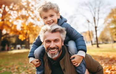 Fototapeta premium A father gives his son a piggyback ride in the park. Portrait of a happy father riding his son on his back on his shoulders and looking up. Cute boy with his father playing outdoors.autumn