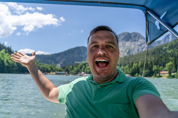The man enthusiastically poses with arms open wide on a boat, smiling against a beautiful lake and...