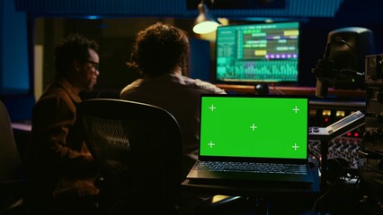 Sound designer and singer look at laptop with isolated display in control room, operating mixing console and volume level buttons to create a hit song. Artist working with audio expert. Camera B.