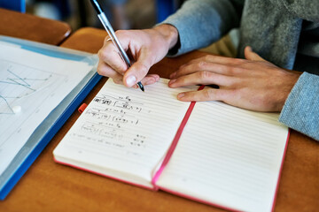 Person, hands and notebook at desk for maths, solving equation or writing solution. Student, book and pen in classroom for higher education, calculus or advanced study at table for learning in school