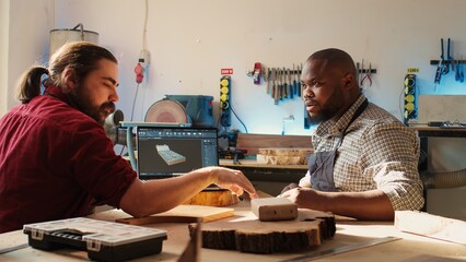 Happy BIPOC cabinetmaker and colleague comparing wood piece with schematic displayed on laptop. Cheerful carpenters in joinery crosschecking wooden object with notebook blueprint