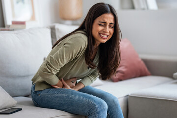 Unhealthy young woman with stomachache leaning on the couch in the living room at home.