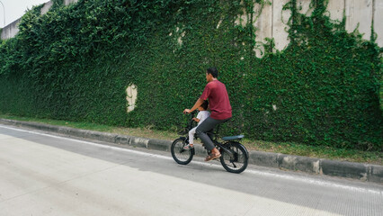 A father and son riding a bicycle on a quiet road, flanked by a tall green ivy wall. A serene urban scene capturing familial joy