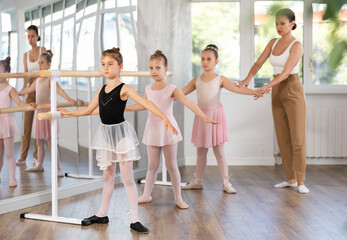 Adult female teacher corrects group of girls dancers standing in second position at ballet barre