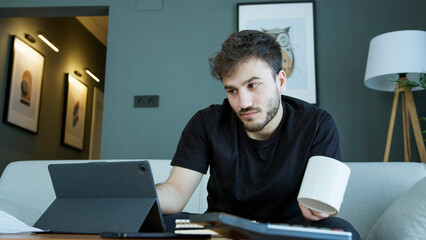 A man sits on a couch, holding a coffee mug while focusing on his tablet. Reviewing important work or studies. 