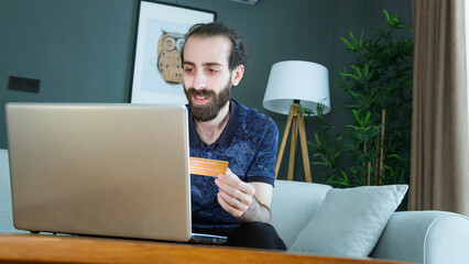 A man with a beard sits on a couch, holding a credit card while shopping online using his laptop.