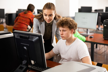 Young female teacher helping focused teenage student studying for exam in college library computer...